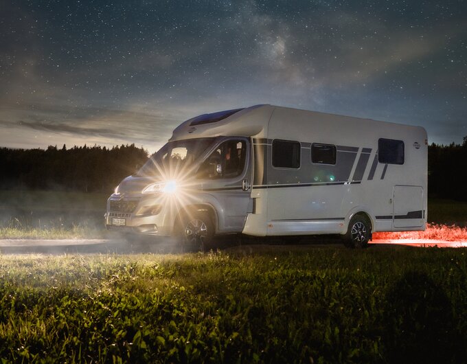 Carado motorhome at night on a meadow at the edge of the forest, side view under a starry sky with the front lights on