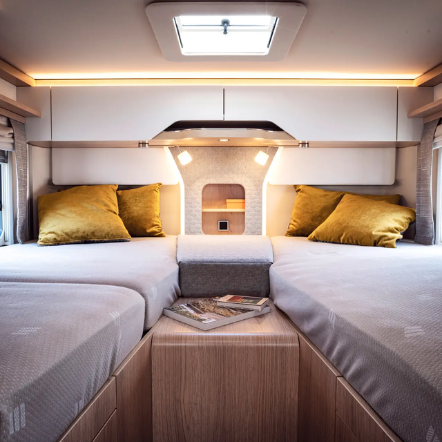 Gezellige slaapruimte achterin een camper met twee eenpersoonsbedden in de lengterichting, verbonden door een extra kussen; twee mosterdkleurige kussens zijn aan elk hoofdeinde.