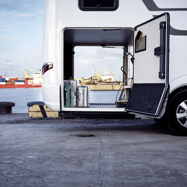 Uitzicht door de garage achter van de HYMER B-ML T-camper, beladen met zilveren koffers, voor de havenfaciliteit met het rode vrachtschip