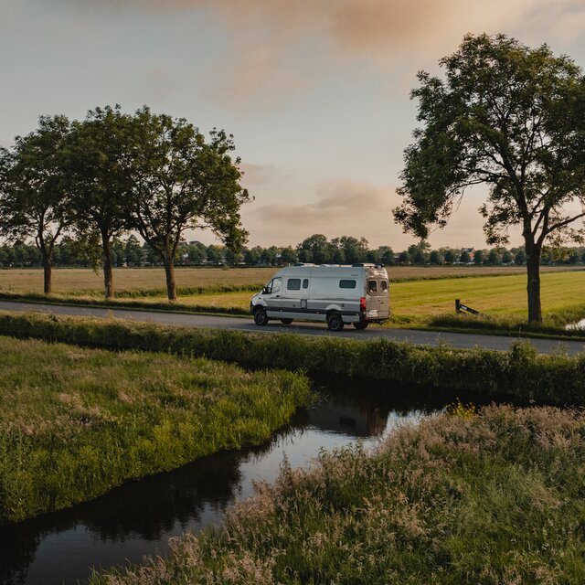 Grey camper van driving on a road, on both sides of the road you can see green fields and small rivers, along the road there are several trees