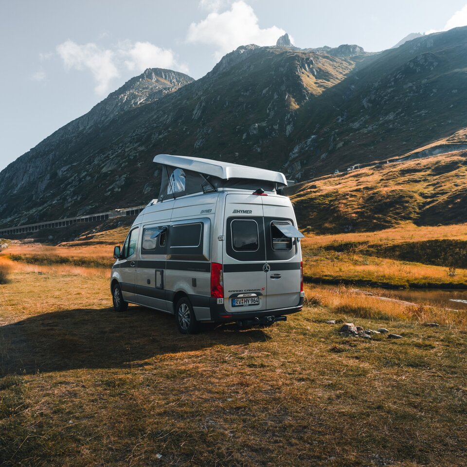 The HYMER Grand Canyon S is seen from slightly behind at an angle. It stands slightly turned in front of a mountain panorama on a meadow.