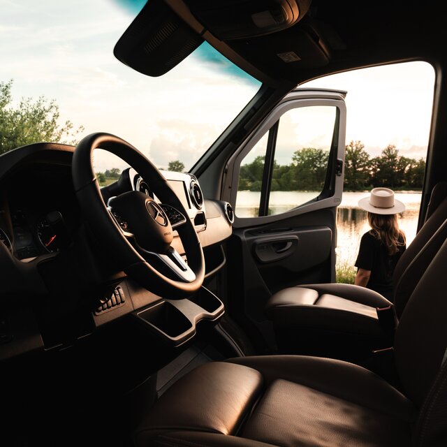 View from the driver's side through the cabin of a Mercedes campervan. Through the open passenger door, you can see a woman wearing a hat looking out at the lake behind.
