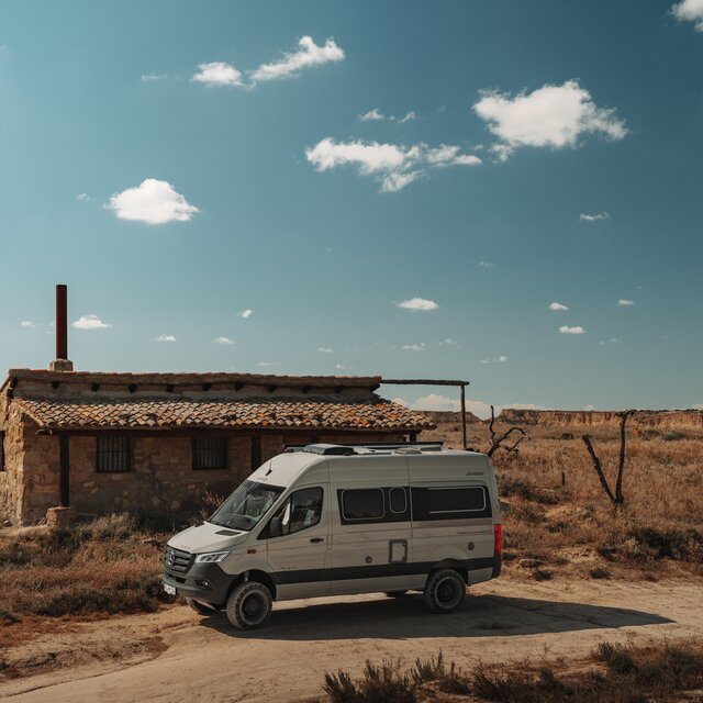 Light grey campervan with black design accents stands sideways in front of a single-storey stone building in a barren, desert-like landscape under a blue sky with very few small clouds