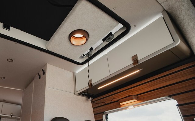 Overhead cabinets above the seating group in a camper van, as well as the open passage at the ceiling leading to the sleeping roof, in this passage the pendant lamp is mounted in a light grey bracket
