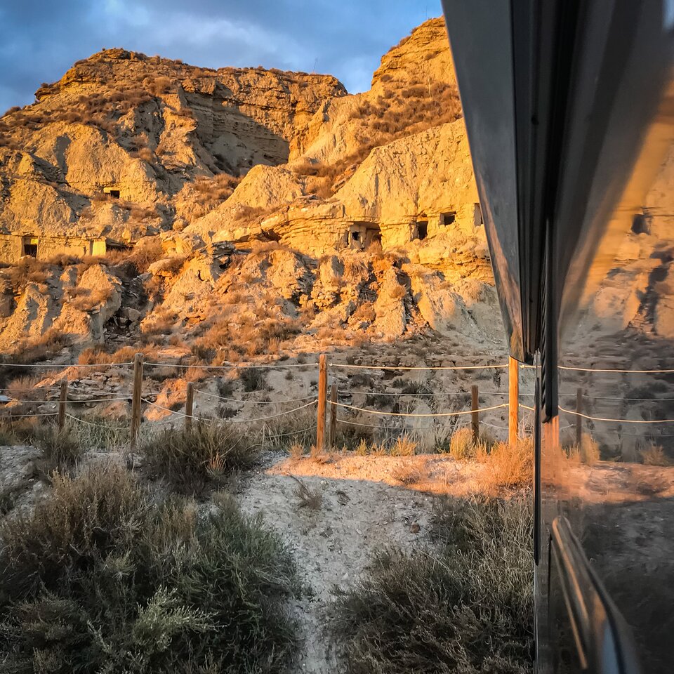 Blick aus dem Wohnmobil in Pyrenäen-Landschaft, seitlich ist die Wand des Reisemobils zu sehen, hinten Büsche und steinige, sonnenbeschienene Hügel.