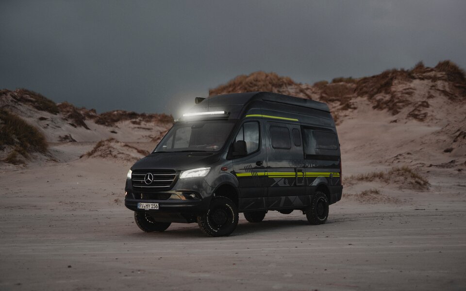Le camping-car tout-terrain HYMER se tient au crépuscule avec phares et barre lumineuse sur une plage de sable avec des dunes en arrière-plan.	